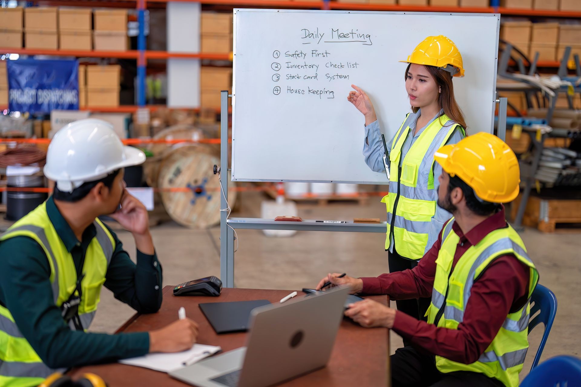 Construction team in safety gear discussing points on a whiteboard during a meeting in a warehouse.