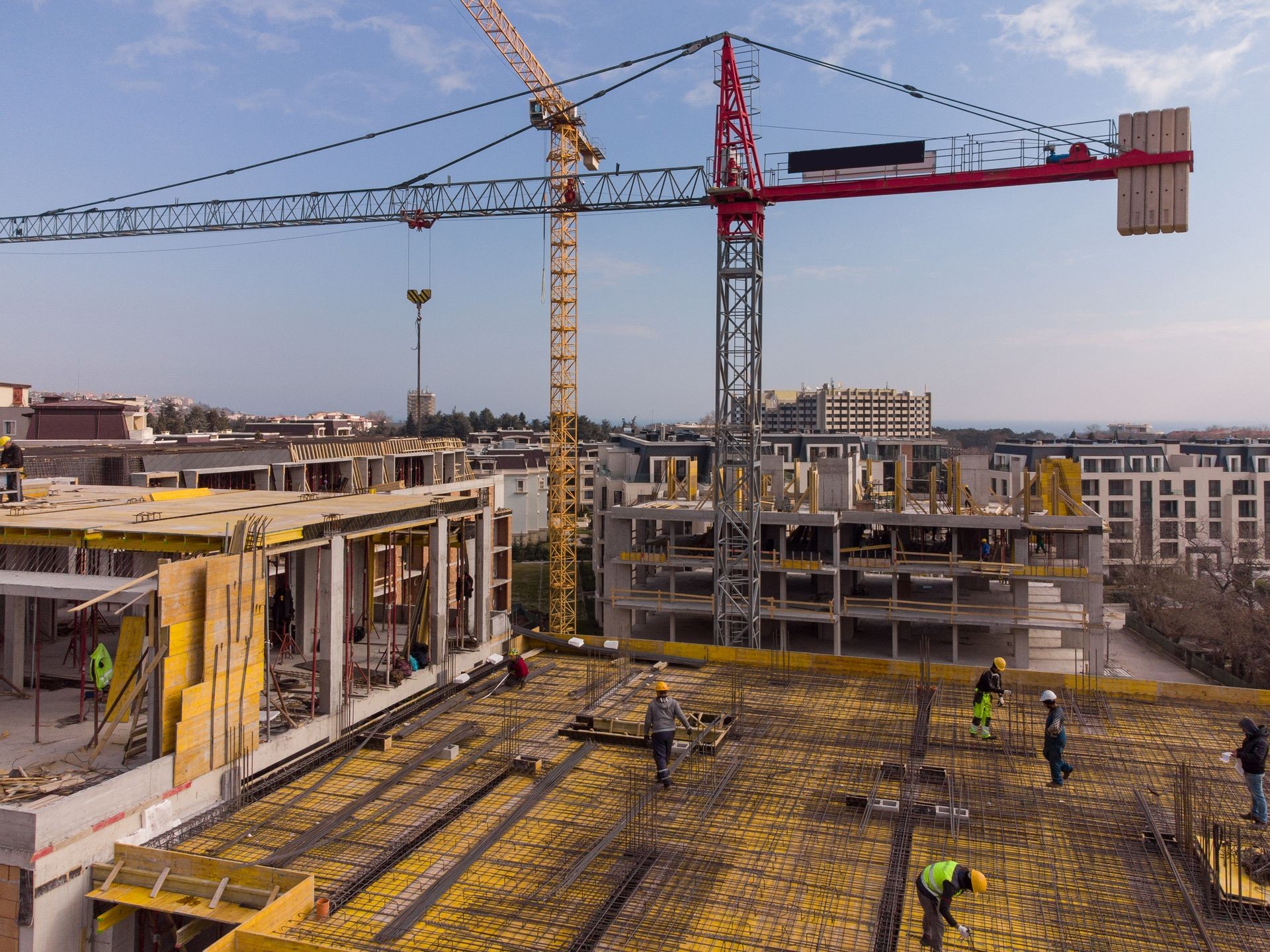 Construction site with workers on a building frame and cranes above, set against a cityscape background.