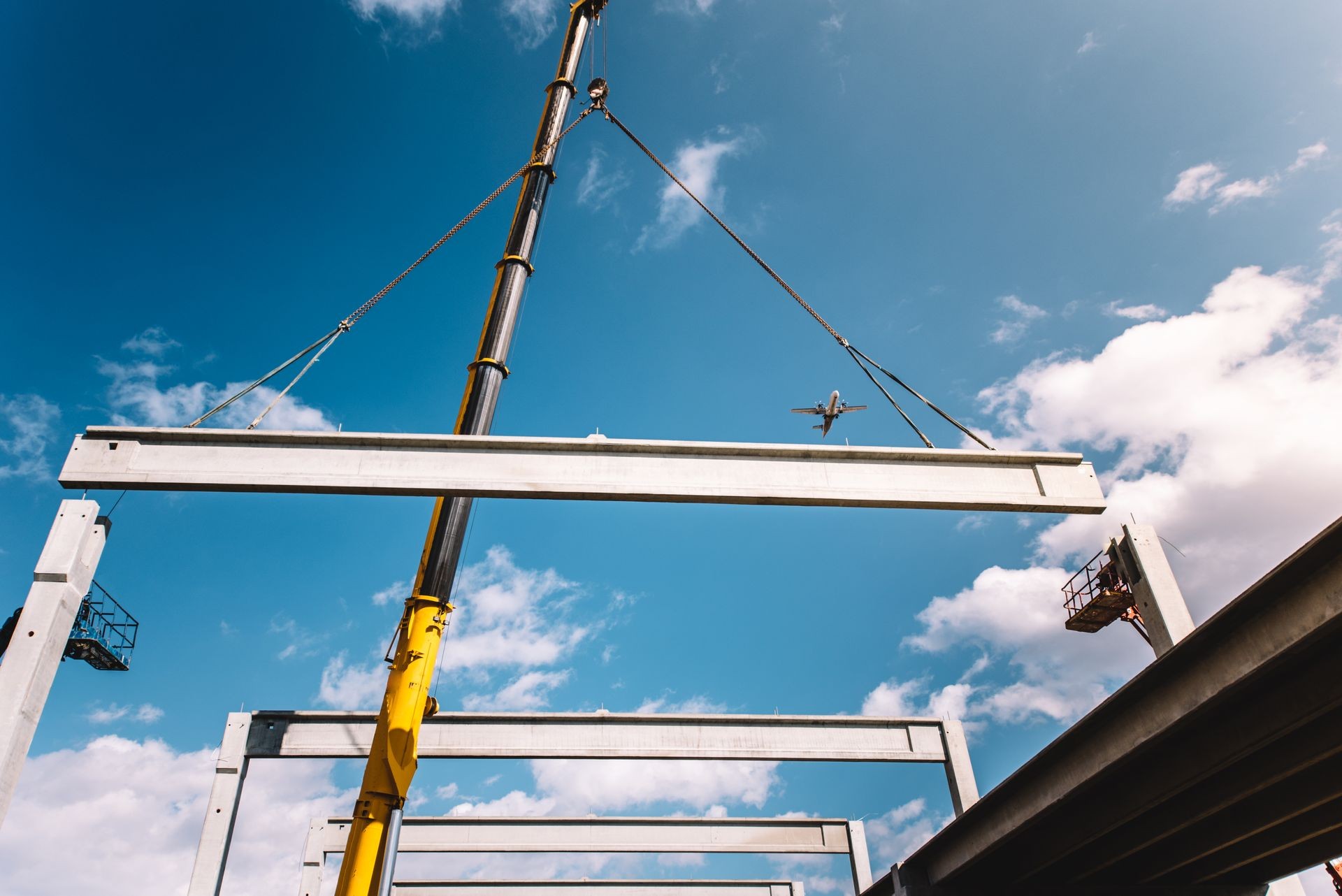 Construction crane lifting a large metal beam against a clear blue sky with an airplane flying overhead.