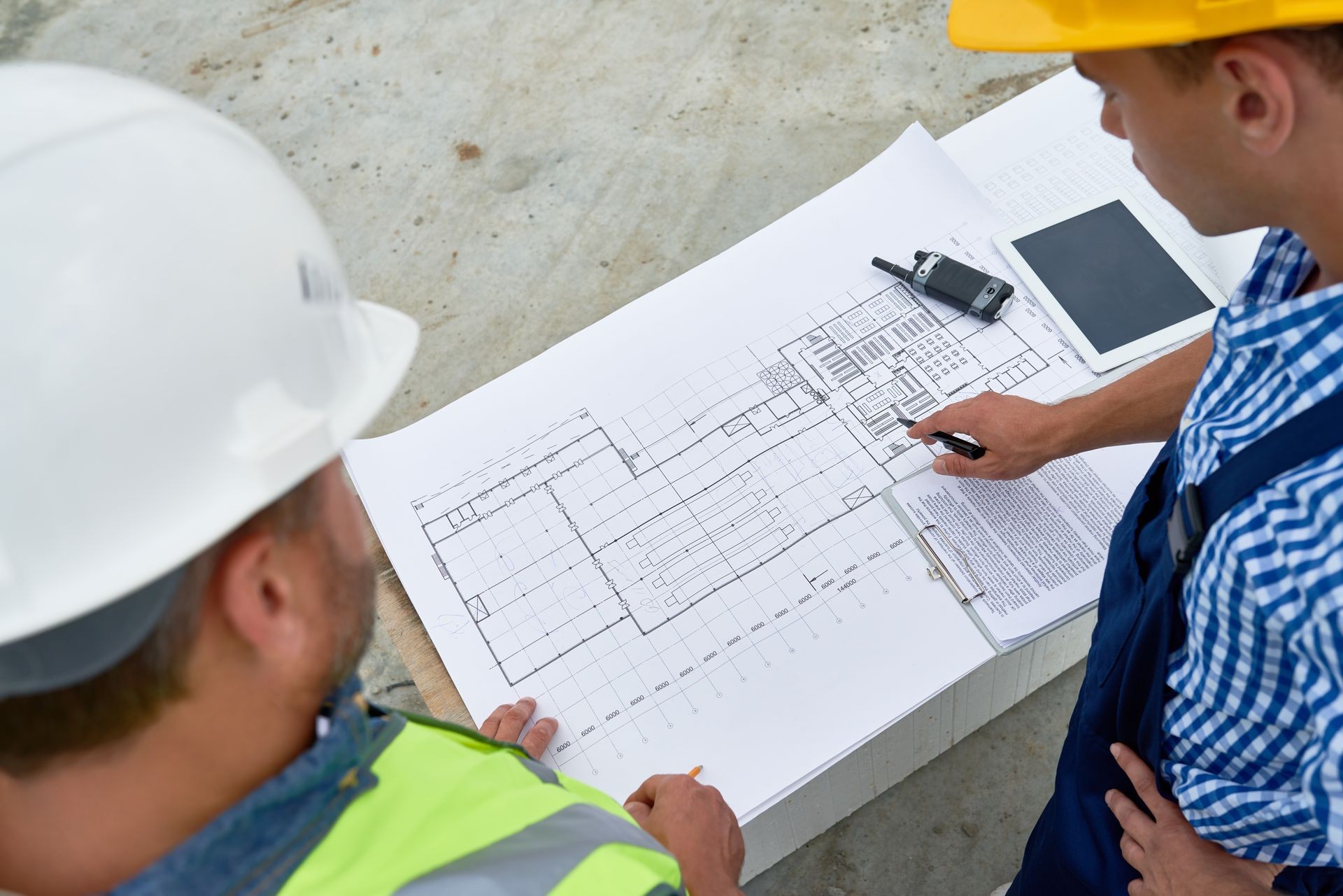 Two construction workers in helmets reviewing architectural blueprints on a table with a tablet and radio.